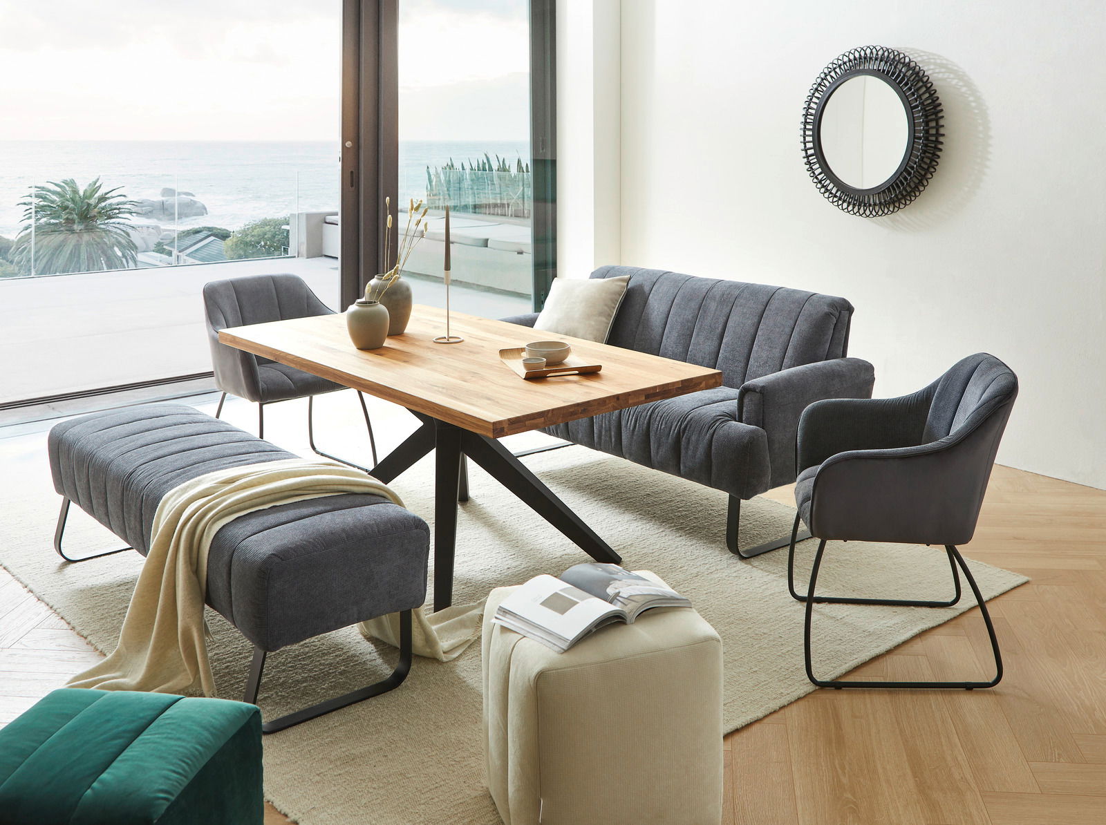 Wohnzimmer mit modernem Esstisch aus Holz, umgeben von grauen Polstermöbeln, darunter ein Armlehnstuhl, eine Sitzbank und ein Sofa. Im Hintergrund ist ein großer Spiegel an der Wand und ein Blick auf das Meer durch die Fenster. Perspektive: seitlich von v