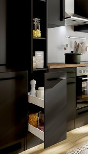 Schuin vooraanzicht van een moderne matzwarte keuken met open apothekerskast en uittreklade, gevuld met voorraadpotten en kruiden, naast een houten werkblad met kookpot.