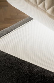 Nahaufnahme eines Boxspringbetts mit grauem Stoffbezug und weißer, strukturierter Matratzenauflage, von oben betrachtet.