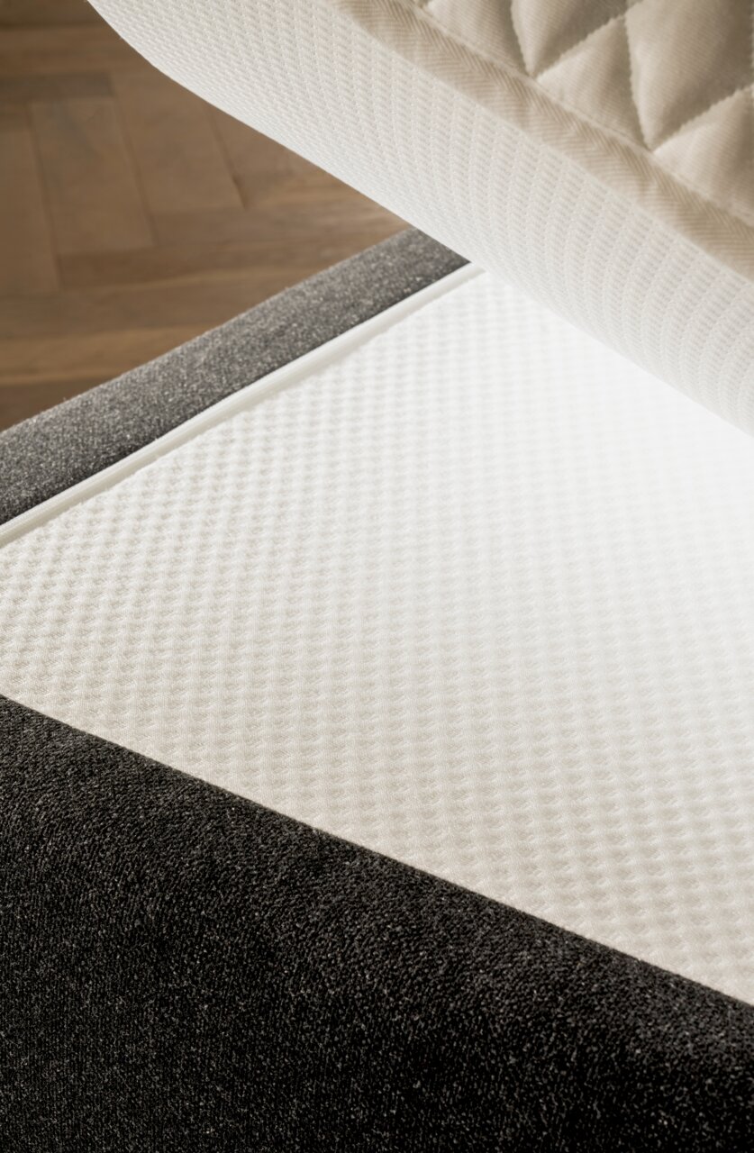 Close-up van een boxspringbed met een grijze stoffen bekleding en een witte, gestructureerde matrashoes, gezien van bovenaf.