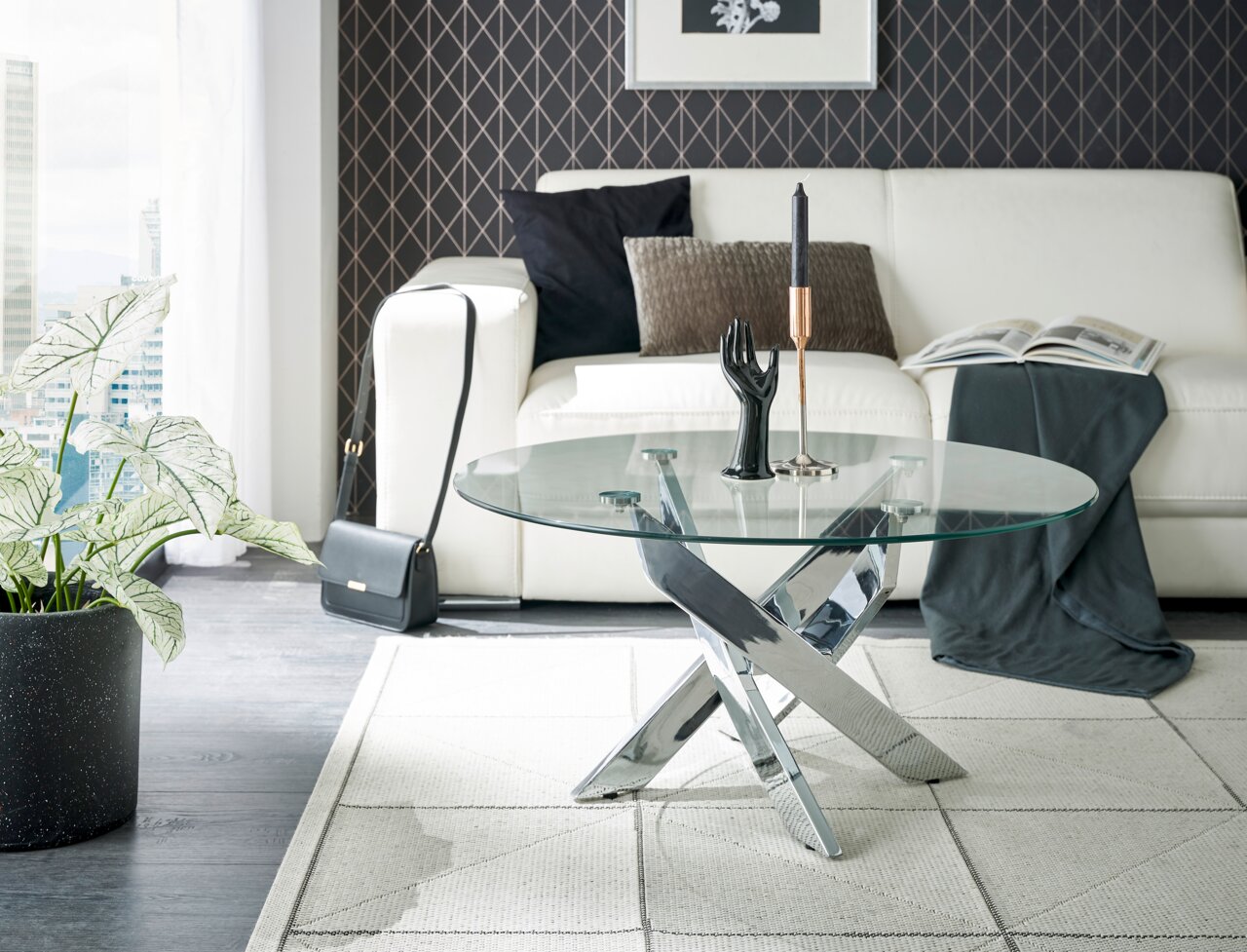 Moderne salontafel met rond glazen blad en verchroomd kruisvormig onderstel, in de woonkamer met een witte bank en decoraties, zijaanzicht