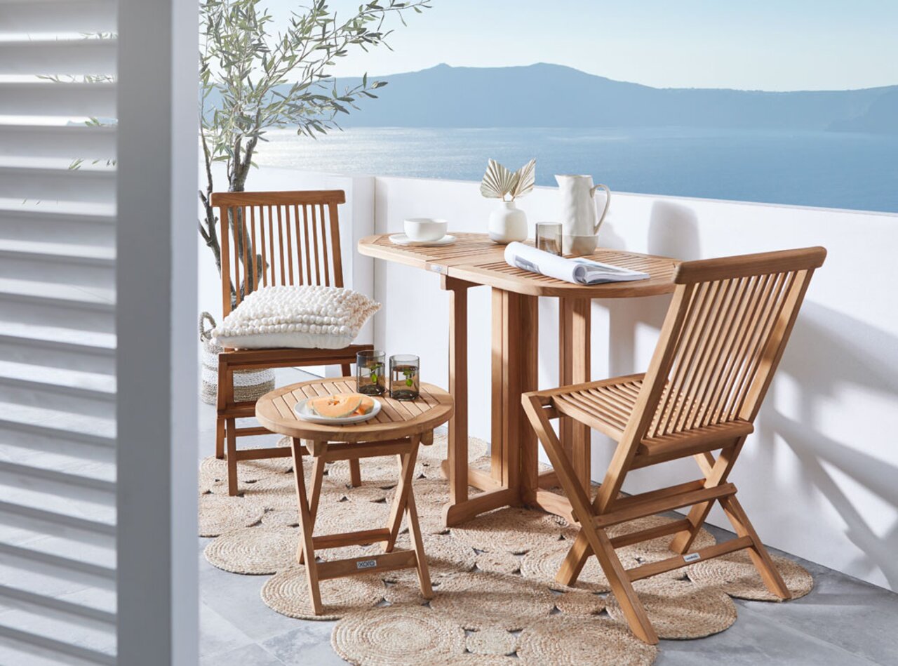 XORA Klapptisch SURABAYA Klapptisch aus Holz auf einem Balkon mit Meerblick, umgeben von zwei Klappstühlen und dekoriert mit Geschirr und Pflanzen, seitliche Perspektive.