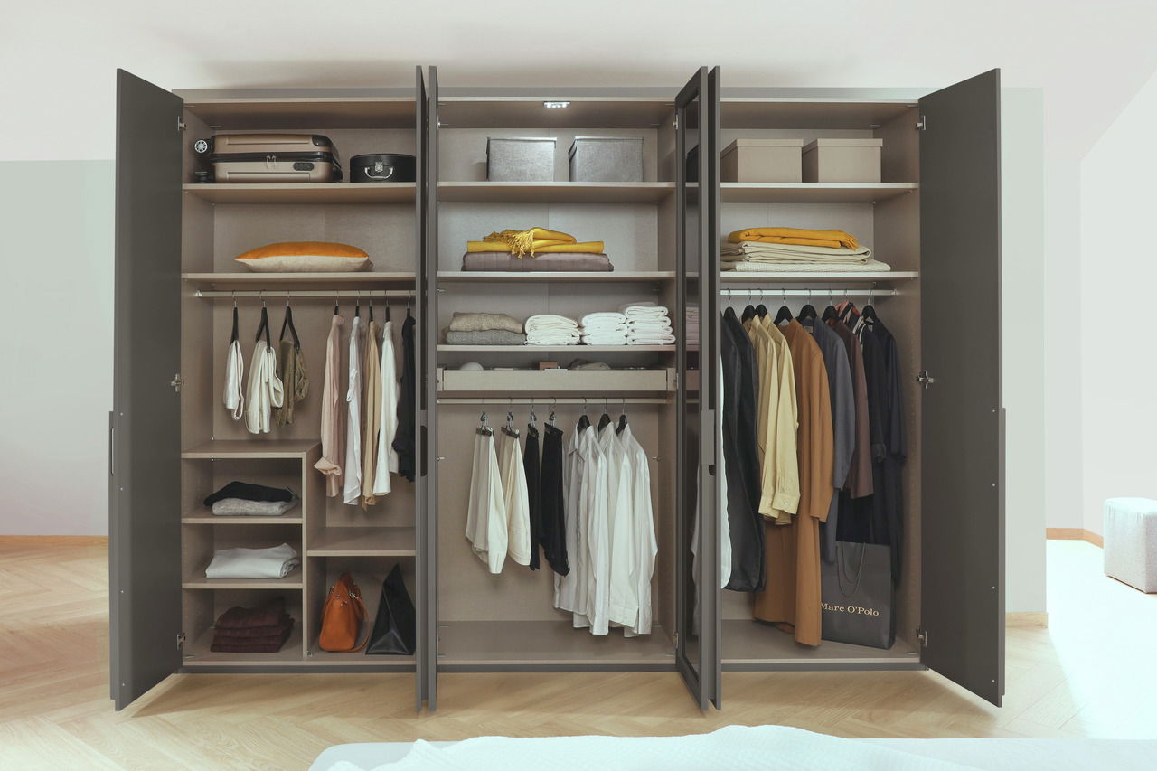 Offener sechstüriger Drehtürenschrank mit verschiedenen Fächern und Kleiderstangen, gefüllt mit Kleidung, Koffern und Boxen, aus frontaler Perspektive.