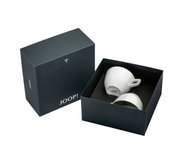 Zwei weiße Tassen in einer eleganten, dunkelgrauen Geschenkbox mit dem Logo 'JOOP!' auf der Unterseite der Tassen, aus der Vogelperspektive fotografiert.