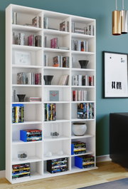 Großes weißes Bücherregal mit vielen offenen Fächern, gefüllt mit Büchern und DVDs sowie Deko-Objekten, im Wohnzimmer vor türkisfarbener Wand; frontale Perspektive mit leichter Ansicht von links.