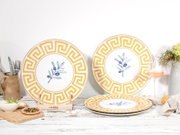 Dekorative Keramikteller im mediterranen Design mit gelbem Mäander-Rand und blauem Olivenzweig-Motiv, auf rustikalem Tisch arrangiert; Perspektive: frontale Aufnahme auf Augenhöhe, leicht seitlich.