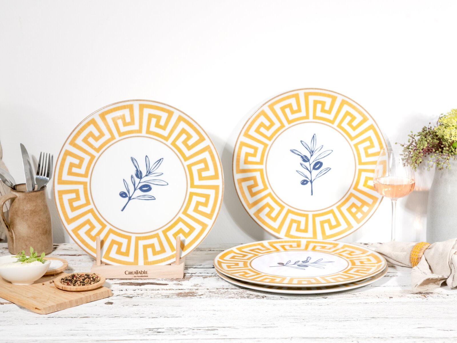 Dekorative Keramikteller im mediterranen Design mit gelbem Mäander-Rand und blauem Olivenzweig-Motiv, auf rustikalem Tisch arrangiert Perspektive: frontale Aufnahme auf Augenhöhe, leicht seitlich.
