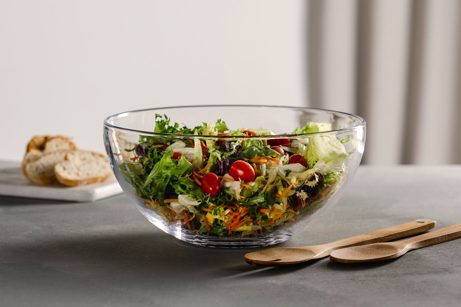 Glasklare Schale mit frischem Salat und Salatbesteck aus Holz, seitliche Perspektive