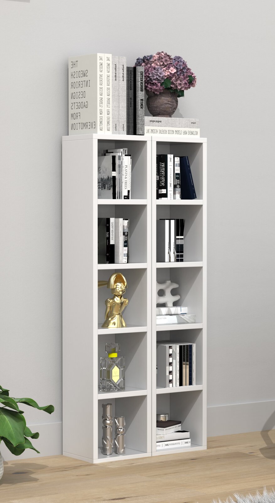 Frontalansicht eines weißen, schmalen Bücherregals mit acht Fächern, dekoriert mit Büchern, goldener Figur, Glasflakon, silbernen Vasen und einer Vase mit rosa Hortensien obenauf, vor hellgrauer Wand auf Holzboden.