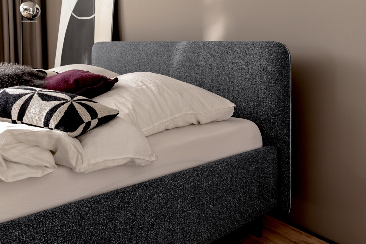 Seitliche Ansicht eines grauen Polsterbetts mit Kopfteil, dekoriert mit Kissen in verschiedenen Farben und Mustern.