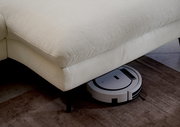 Nahaufnahme einer cremefarbenen Longchairkombination mit einem Saugroboter, der darunter hervorschaut. Perspektive von der Seite.