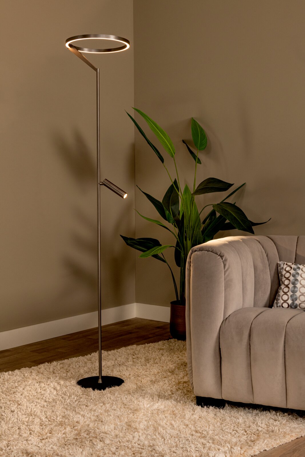 Moderne LED-Stehleuchte aus Metall mit ringförmigem Leuchtkopf und zusätzlichem Lesearm, eingeschaltet neben Sofa und Zimmerpflanze im Wohnzimmer, perspektivisch in leichter Seitenansicht aufgenommen.