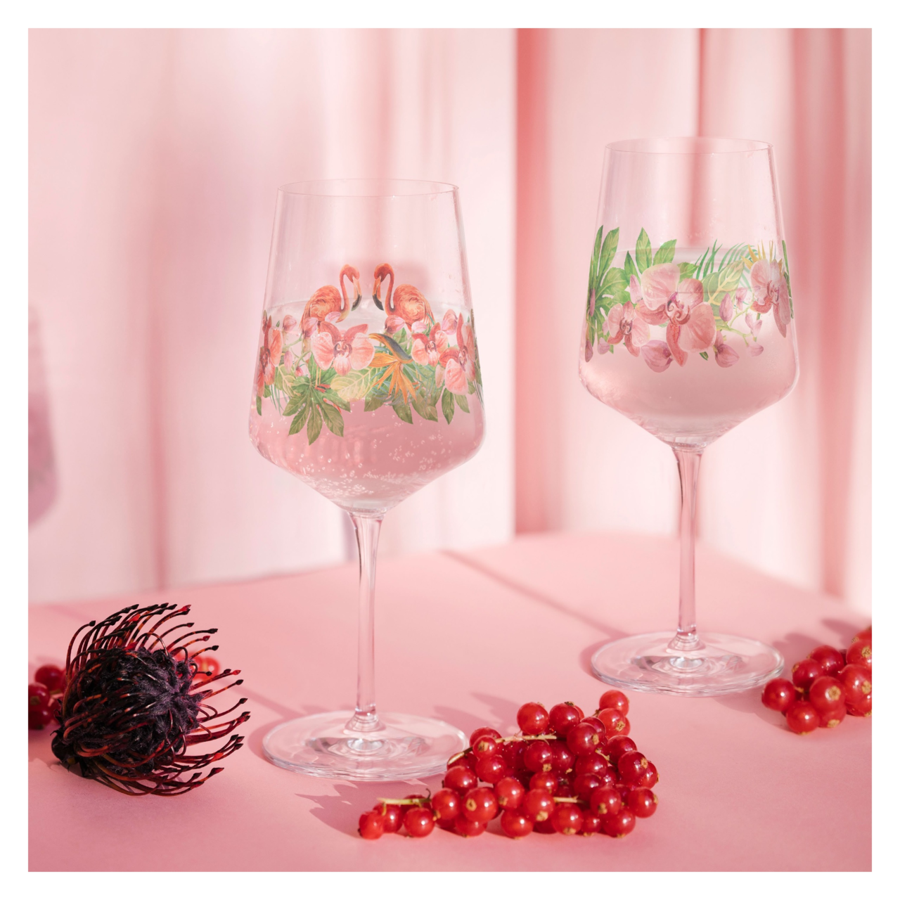 Zwei Weingläser mit floralen und Flamingo-Motiven, gefüllt mit einem rosa Getränk, auf einem rosa Hintergrund mit roten Beeren im Vordergrund. Frontalansicht.