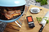 Digitales Bratenthermometer neben einem Grill mit Fleisch, aus der Vogelperspektive aufgenommen.