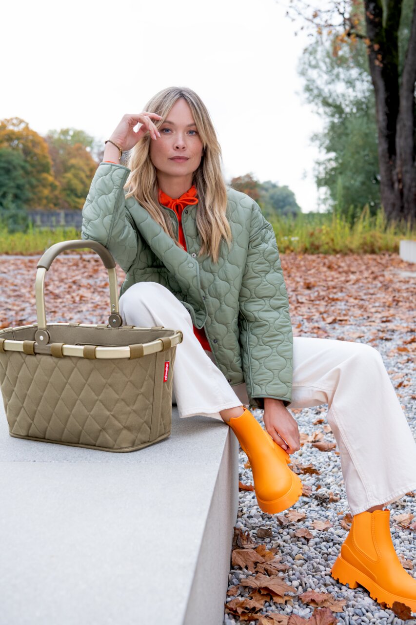 Carrybag Rhombus Olive in een herfstachtige omgeving, van voren gefotografeerd, naast een persoon met een olijfgroen jasje en oranje laarzen.