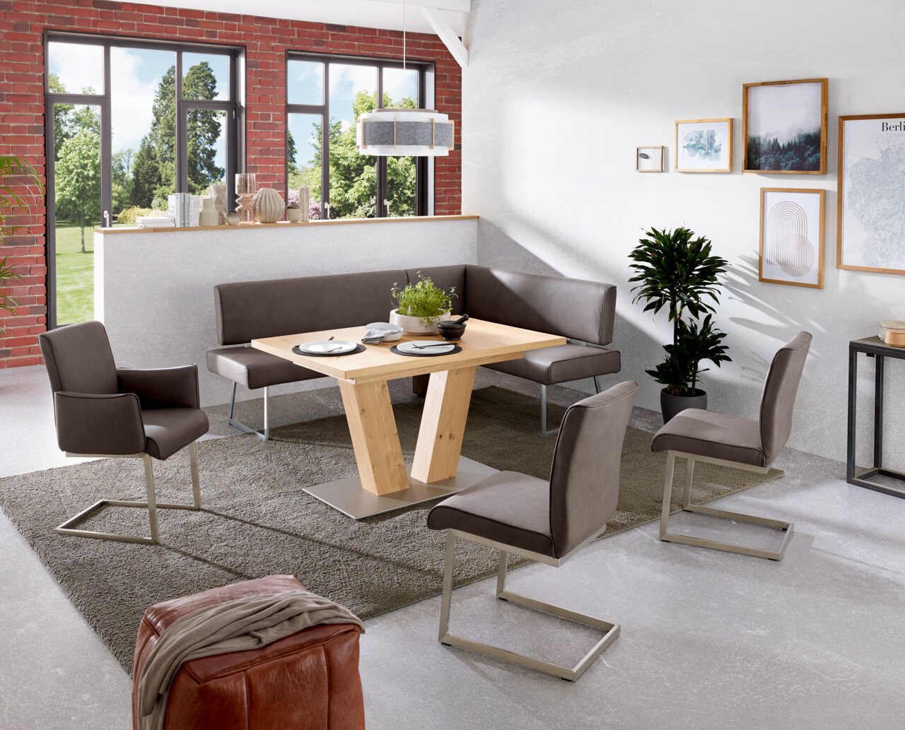 Moderner Esstisch aus Holz mit V-förmigem Gestell, umgeben von grauen Stühlen und einer Eckbank in einem hellen Raum mit großen Fenstern. Perspektive: seitlich von oben.