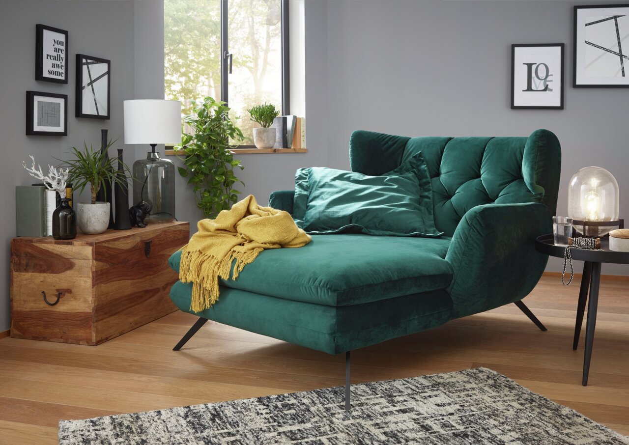 Grünes Longseat mit Samtbezug in einem modernen Wohnzimmer, seitliche Perspektive, dekoriert mit gelber Decke und Kissen.