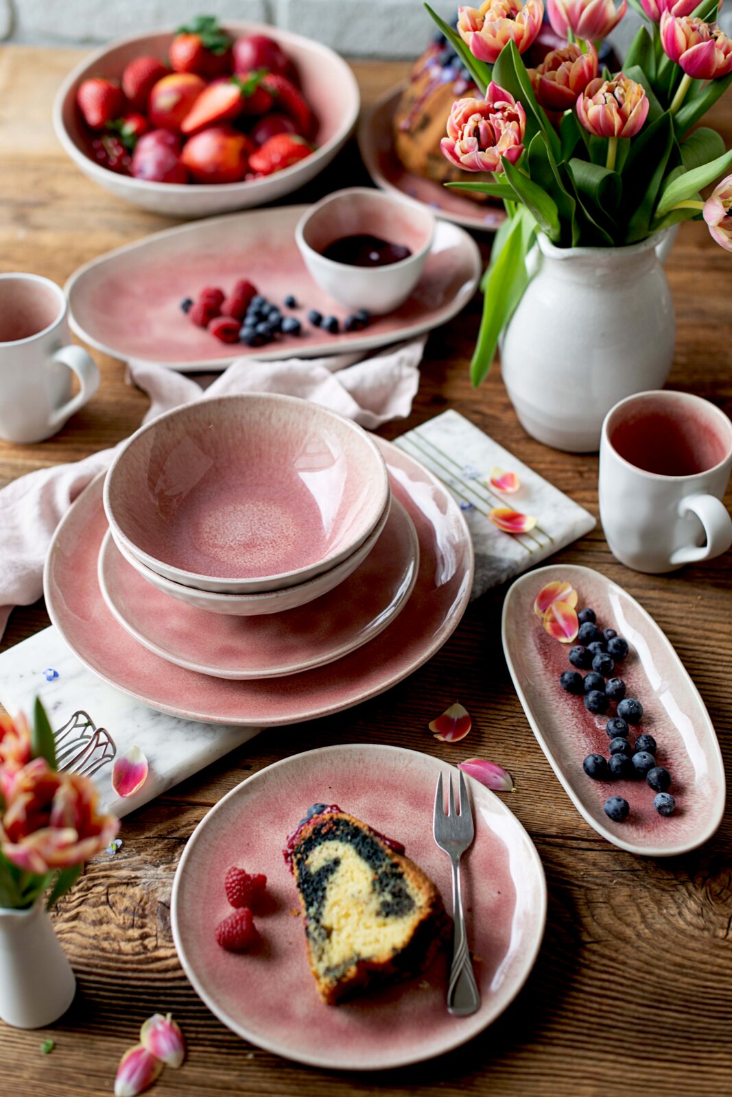 Rosafarbenes Keramikgeschirr mit Tellern, Schalen, Tassen und Servierplatten, stilvoll arrangiert mit Beeren, Marmorkuchen und Tulpen auf einem rustikalen Holztisch, aus schräger Vogelperspektive.