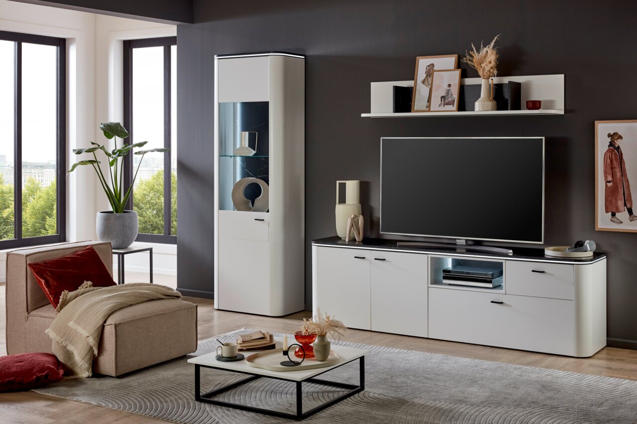 Zurbrüggen Vitrine WAVE Moderne Wohnzimmeransicht mit einer weißen Vitrine, die von vorne gezeigt wird, neben einem großen Fernseher auf einem weißen TV-Schrank. Ein bequemer Sessel mit Kissen und ein Couchtisch mit Dekorationen runden das stilvolle Ambiente ab.