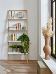 Modernes Leiterregal aus hellem Holz mit weißen Ablagen, dekoriert mit Büchern, Pflanzen und Uhr, in heller Wohnraumszene; Perspektive: frontale Ansicht auf Augenhöhe.