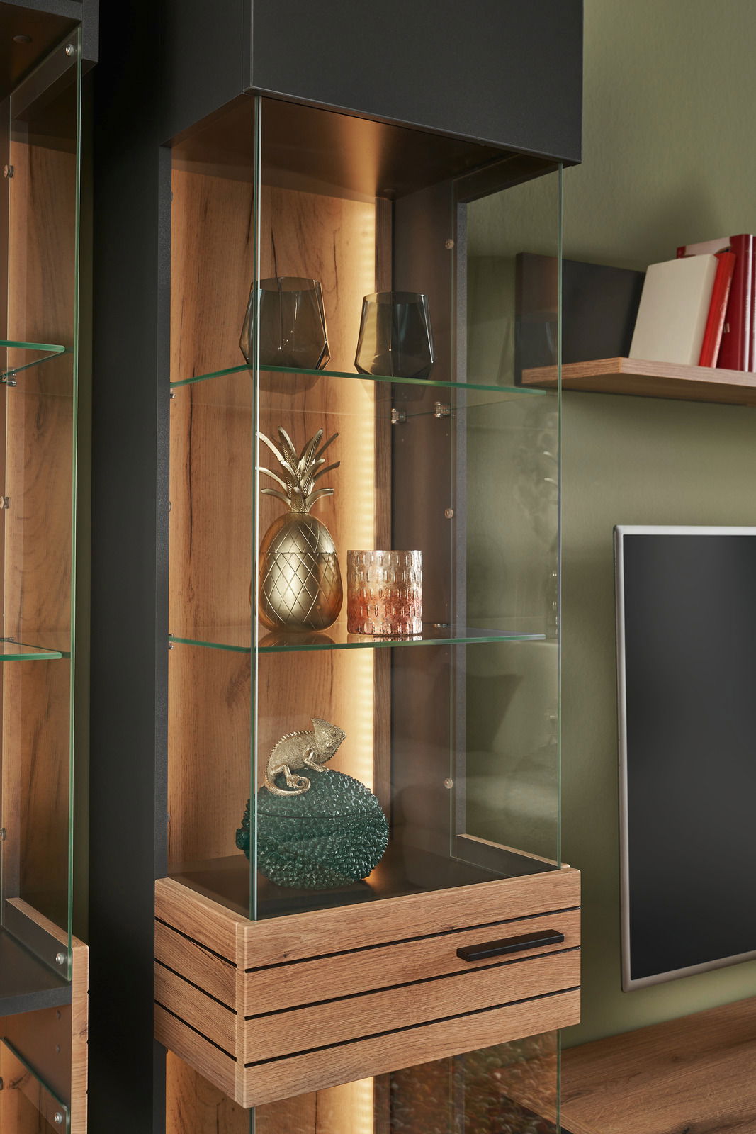 Nahaufnahme einer modernen Vitrine mit Glasregalen, dekoriert mit Gläsern, einer goldenen Ananas und einem kleinen Deko-Chamäleon. Die Perspektive zeigt die Vitrine von vorne leicht seitlich.