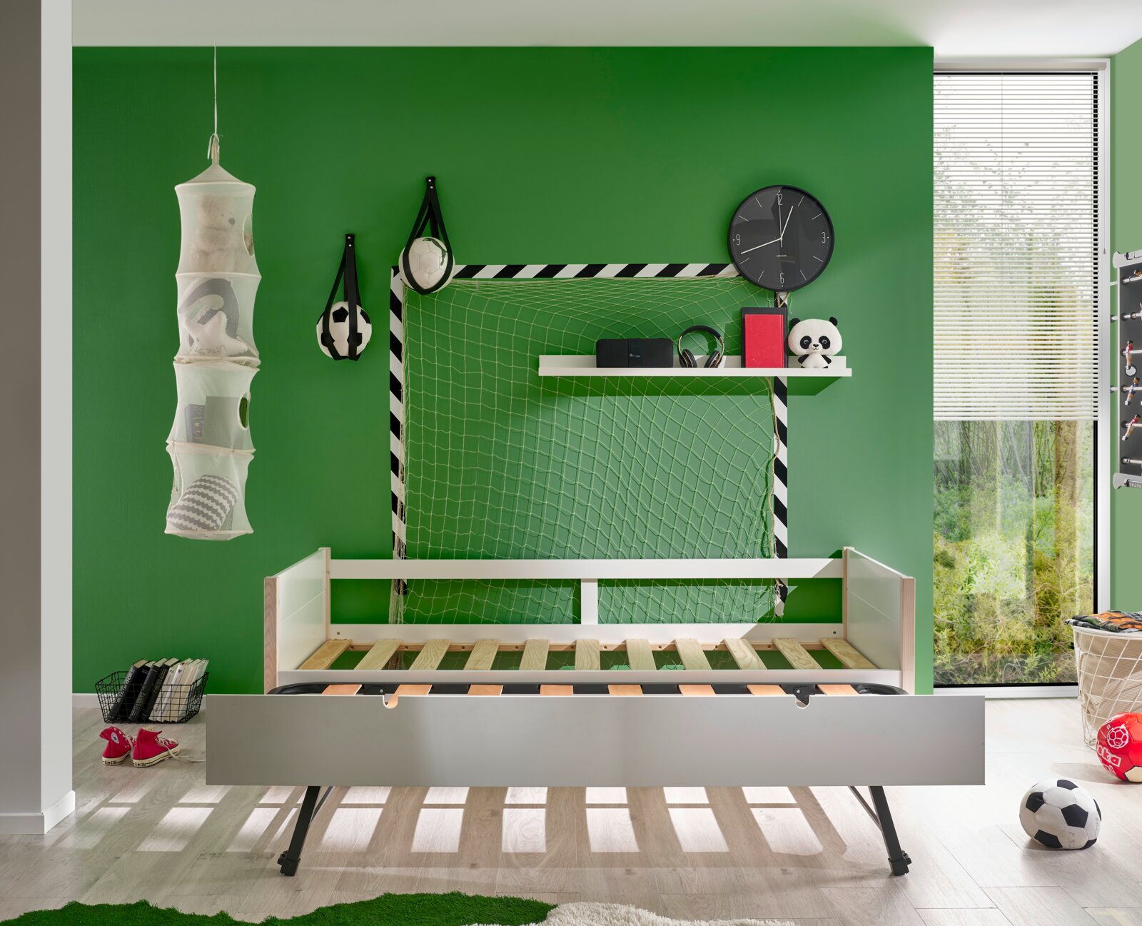 Jeugdbed in een sportief ingerichte kinderkamer met een groen geschilderde muur, voetbalversieringen en een groot raam. Perspectief: vooraanzicht van het bed.