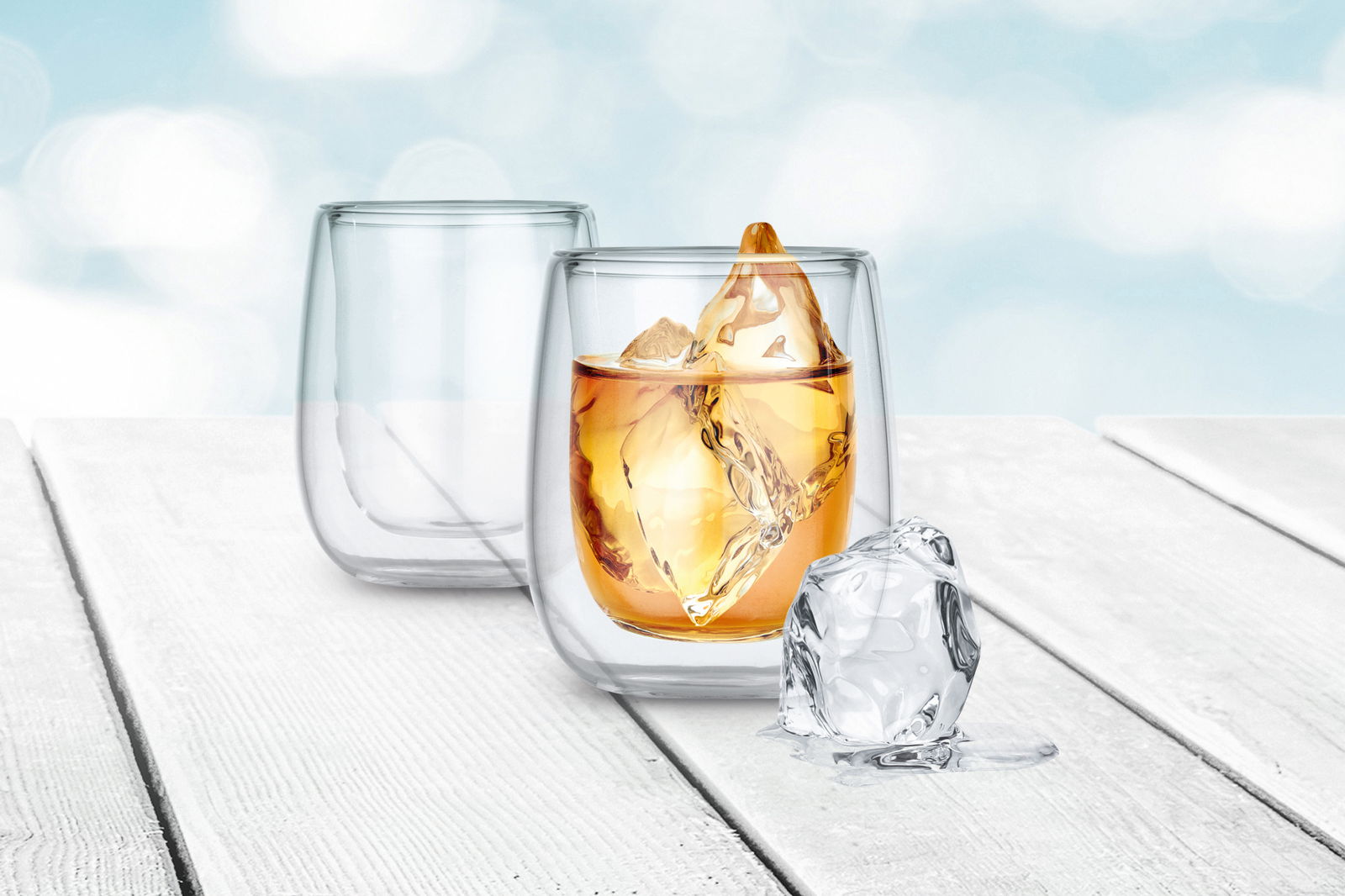 Zwei doppelwandige Thermogläser auf einem Holztisch, eines gefüllt mit einem Getränk und Eiswürfeln, aus seitlicher Perspektive fotografiert.