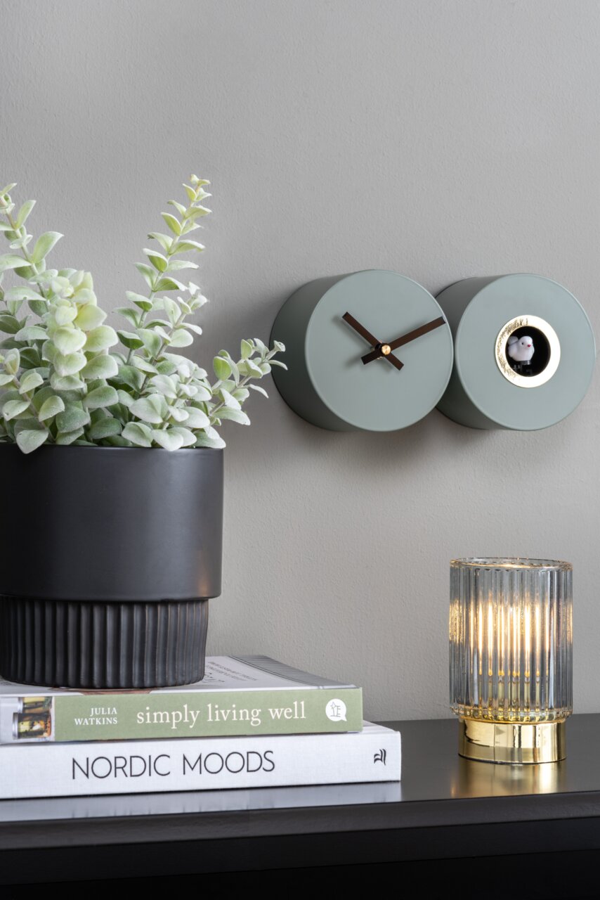 Wandklok in mat jungle-groen met koekoek, zijaanzicht, naast een plant en een lamp op een plank.