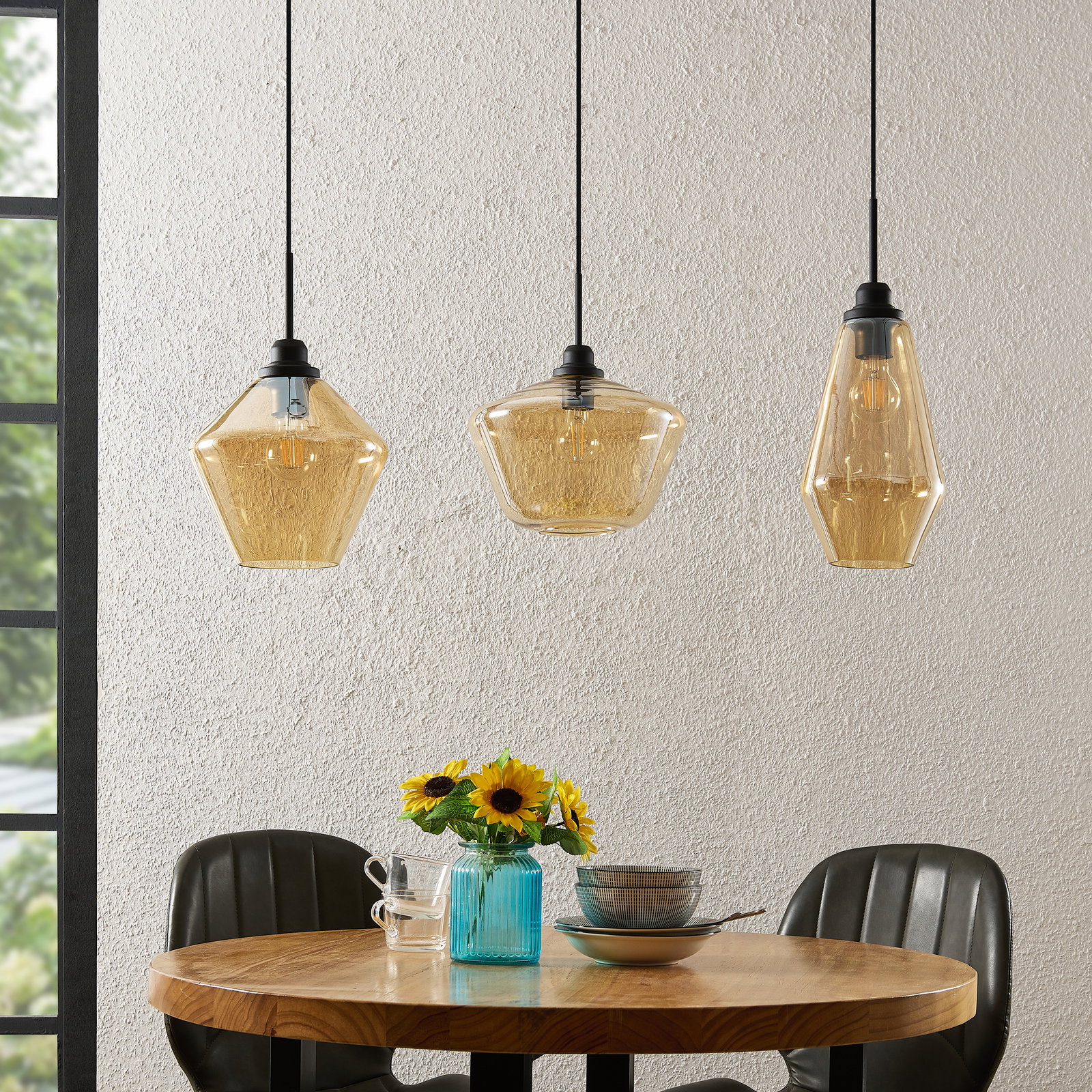 Drei moderne Hängeleuchten aus Glas in verschiedenen geometrischen Formen, hängend über einem runden Holztisch mit Blumen und Geschirr, seitliche Perspektive.