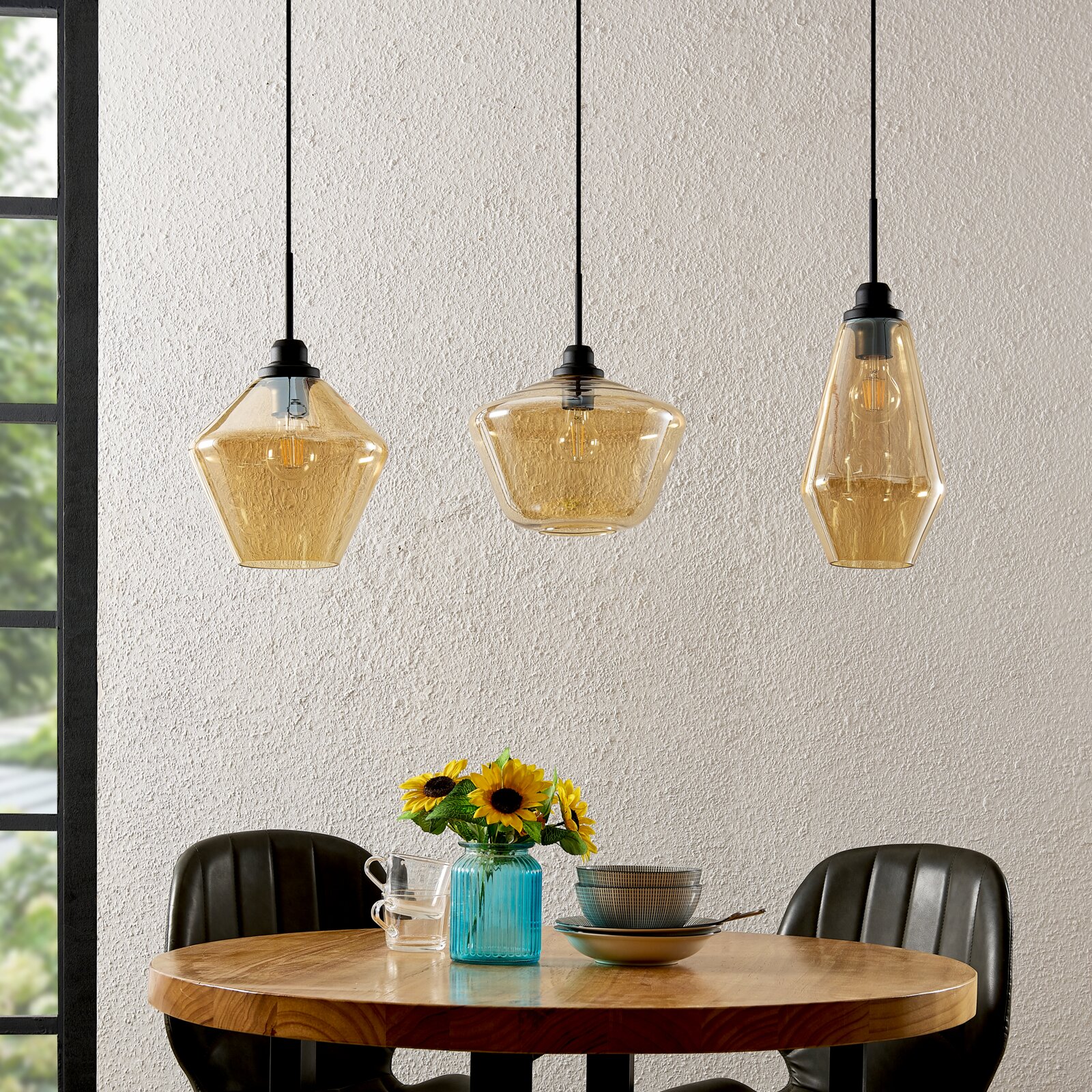Drei moderne Hängeleuchten aus Glas in verschiedenen geometrischen Formen, hängend über einem runden Holztisch mit Blumen und Geschirr, seitliche Perspektive.