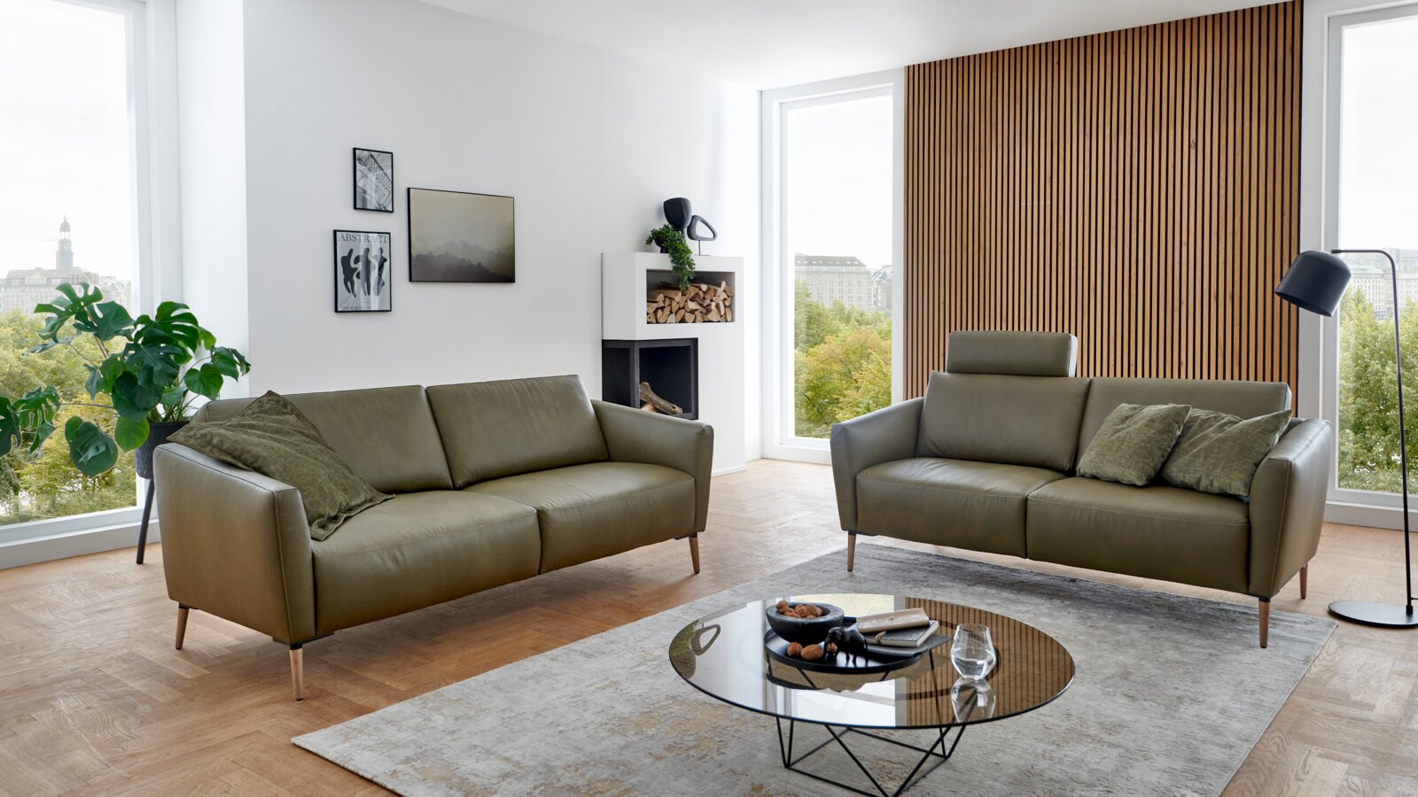 Valnatura Sofa 3er PERLA Zwei grüne Sofas in einem modernen Wohnzimmer mit großen Fenstern, Holzboden und stilvoller Dekoration, seitliche Perspektive.