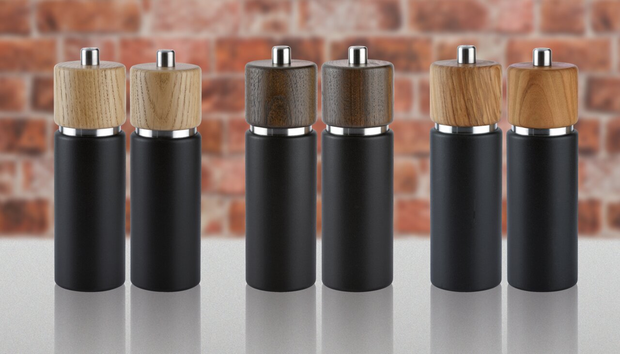 Sechs Salzmühlen in verschiedenen Holzarten, frontal auf einer glänzenden Oberfläche vor einer Ziegelwand präsentiert.