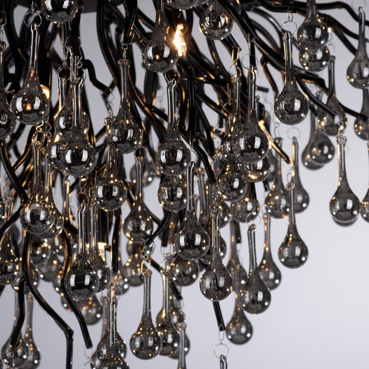 Close-up van een plafondlamp met druppelvormige glazen elementen die aan zwarte metalen armen hangen. De foto is van onderaf genomen.