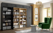 Moderne Wohnzimmeransicht mit einem großen, grauen Mehrzweckschrank und einem offenen Holzregal, gefüllt mit Büchern und Aufbewahrungsboxen. Ein grüner Sessel und ein kleiner runder Tisch stehen vor einem großen Fenster mit Vorhängen. Perspektive: Frontal