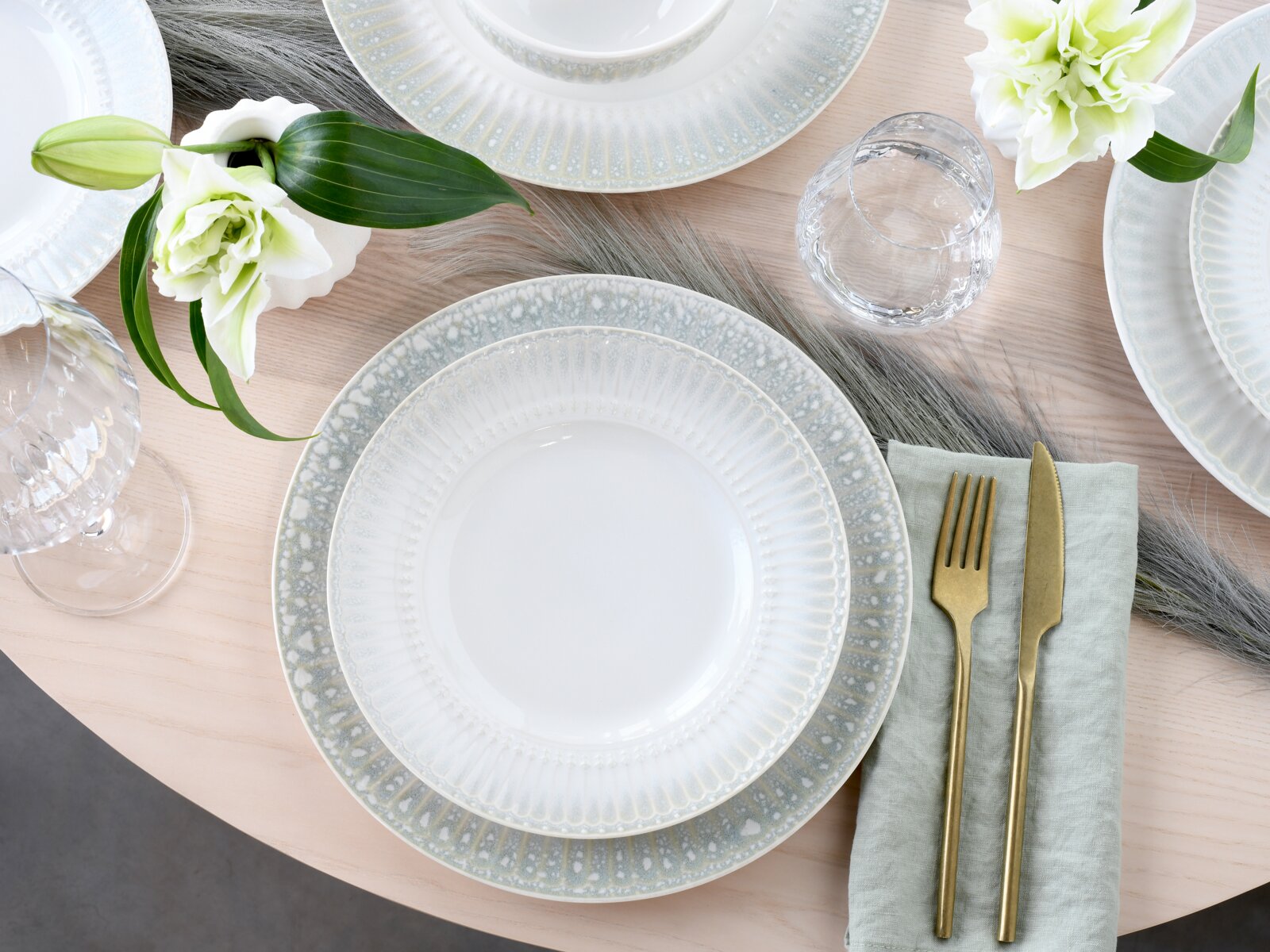 Elegantes Tafelgedeck mit weiß-grau gesprenkelten Keramiktellern, Glas und goldfarbenem Besteck auf hellem Holztisch mit Blumen, Draufsicht (Top-Down).