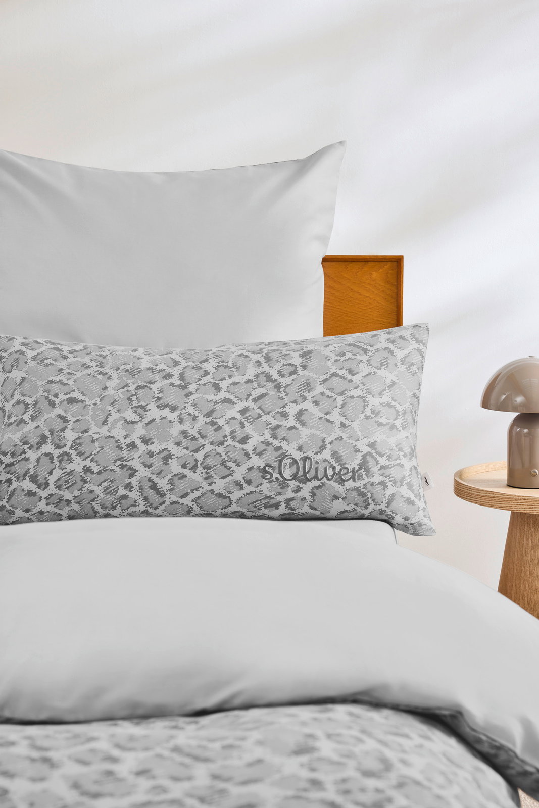 Bettwäsche-Set in Grau mit Leopardenmuster, bestehend aus einem Kissenbezug und einem Bettbezug, auf einem Bett mit Holzrahmen und einem Nachttisch mit Lampe, aus der Perspektive von vorne.
