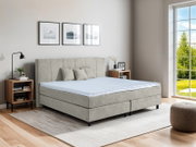 Modernes Boxspringbett in einem hellen Schlafzimmer, seitliche Perspektive mit grauem Stoffbezug, zwei Kissen und einem Nachttisch.