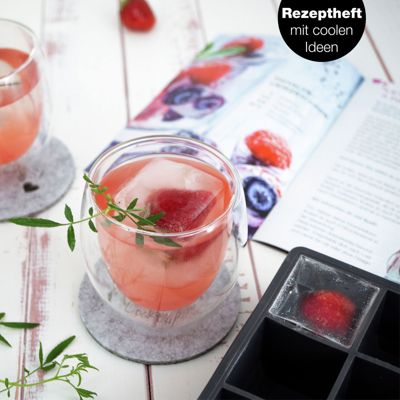 Nahaufnahme eines Getränks mit Eiswürfeln und Erdbeeren in einem doppelwandigen Glas, daneben eine Eiswürfelform und ein Rezeptheft.