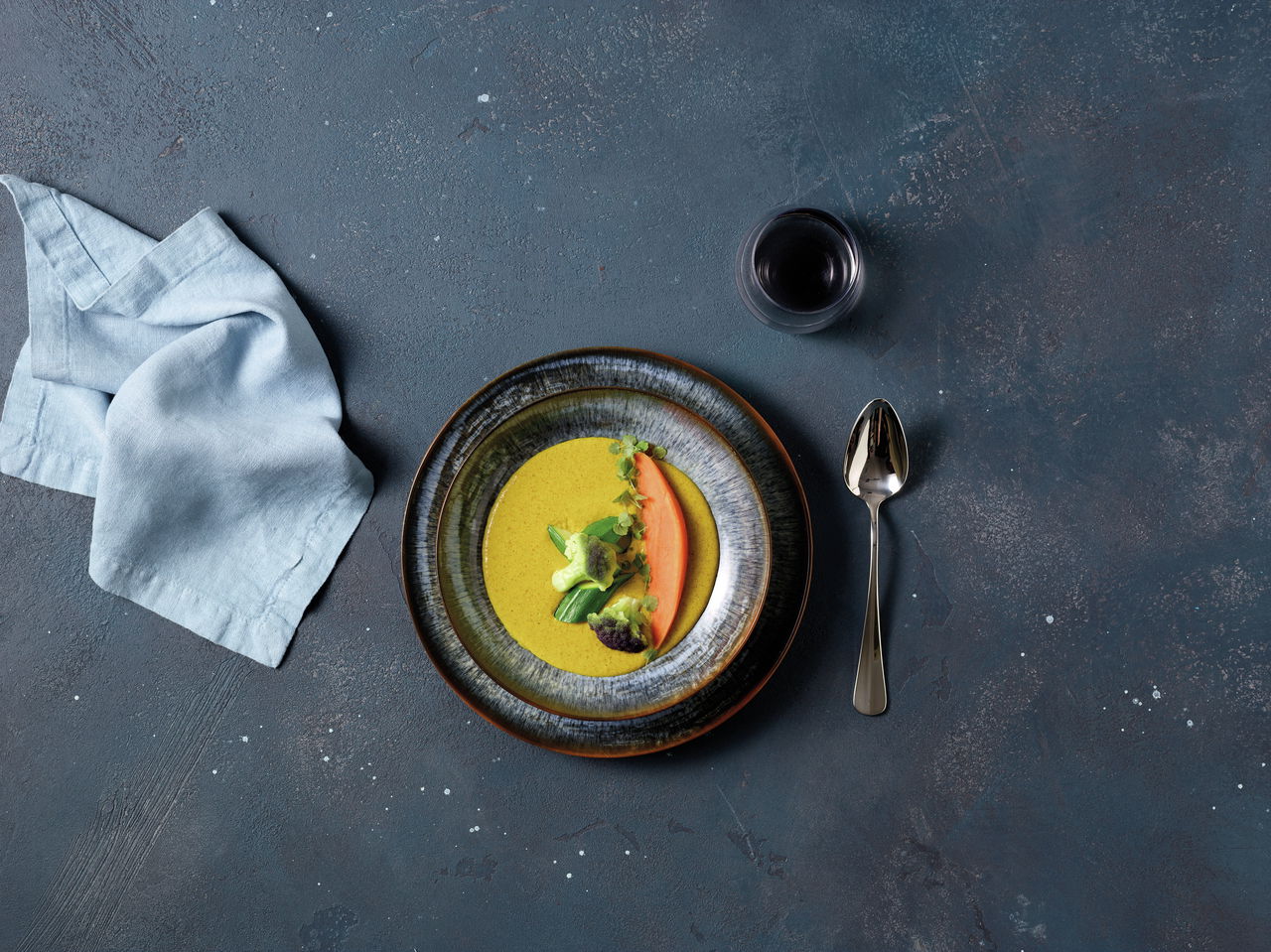 Arthur Krupp Tellerset 12-tlg. Galaxy Neptune Vogelperspektive auf einen Teller aus dem Tellerset Galaxy Neptune mit dekorativ angerichteter Suppe und Gemüse, daneben ein Löffel, ein Glas Rotwein und eine Stoffserviette.