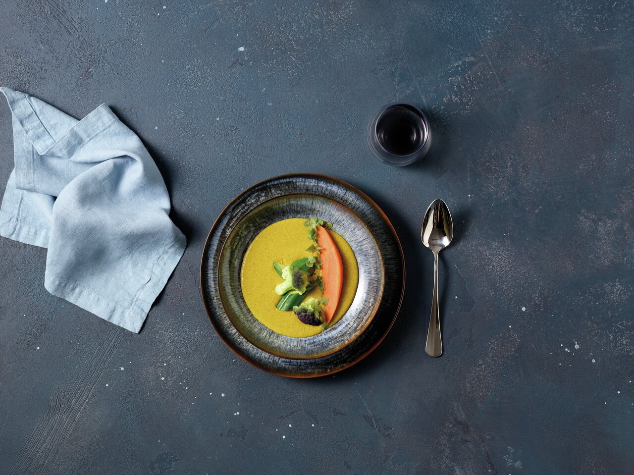 Vogelperspektive auf einen Teller aus dem Tellerset Galaxy Neptune mit dekorativ angerichteter Suppe und Gemüse, daneben ein Löffel, ein Glas Rotwein und eine Stoffserviette.