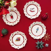 Vogelperspektive auf ein festliches Set von vier Jubiläumstellern mit weihnachtlichen Motiven wie Weihnachtsmann, Schaukelpferd, Nussknacker und Spielzeugzug, umgeben von dekorativen Weihnachtskugeln und Blumen auf rotem Hintergrund.