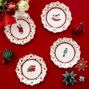 Vogelperspektive auf ein festliches Set von vier Jubiläumstellern mit weihnachtlichen Motiven wie Weihnachtsmann, Schaukelpferd, Nussknacker und Spielzeugzug, umgeben von dekorativen Weihnachtskugeln und Blumen auf rotem Hintergrund.
