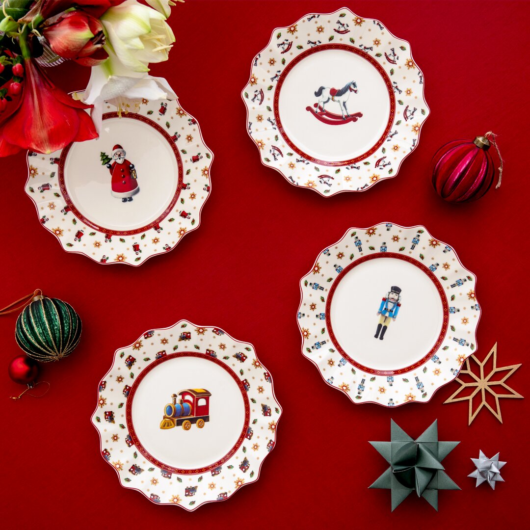 Vogelperspektive auf ein festliches Set von vier Jubiläumstellern mit weihnachtlichen Motiven wie Weihnachtsmann, Schaukelpferd, Nussknacker und Spielzeugzug, umgeben von dekorativen Weihnachtskugeln und Blumen auf rotem Hintergrund.