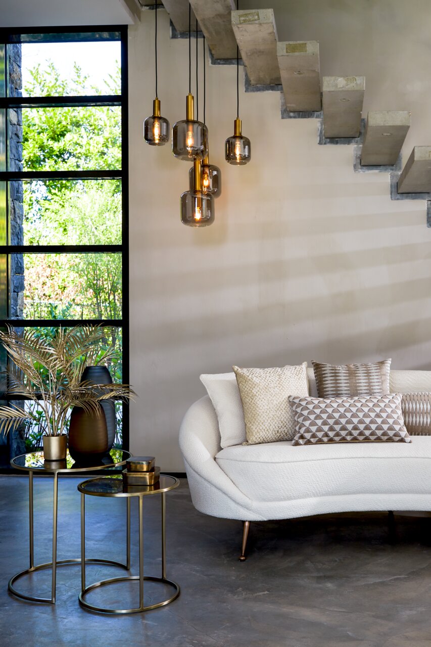 Twee elegante bijzettafeltjes in goud met glazen bladen, versierd met een vaas en planten, in een moderne woonkamer met een witte bank en hanglampen. Aanzicht van voren.