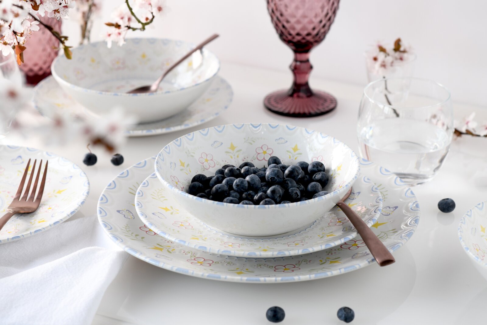 Pastellfarbenes Geschirrset mit floralem Muster: Schale mit Blaubeeren auf Teller, daneben Besteck und Gläser auf weißem Tisch, fotografiert in leicht schräger Seitenansicht