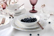 Pastellfarbenes Geschirrset mit floralem Muster: Schale mit Blaubeeren auf Teller, daneben Besteck und Gläser auf weißem Tisch, fotografiert in leicht schräger Seitenansicht
