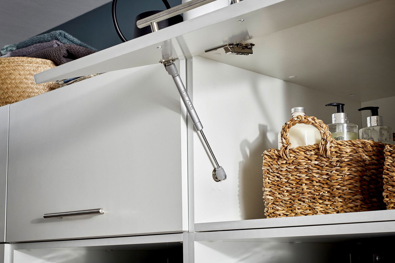 Nahaufnahme eines geöffneten weißen Aufsatzschranks mit einem Gasdruckdämpfer, geflochtenen Körben und Pflegeprodukten. Perspektive von schräg unten.