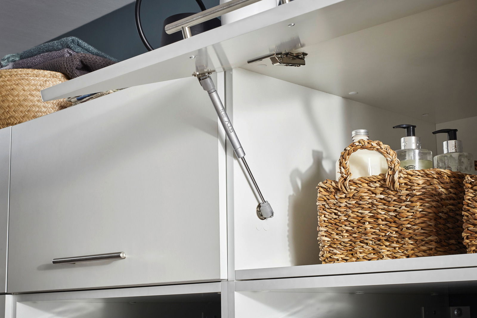Nahaufnahme eines geöffneten weißen Aufsatzschranks mit einem Gasdruckdämpfer, geflochtenen Körben und Pflegeprodukten. Perspektive von schräg unten.