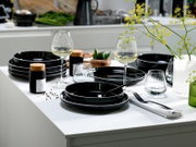 Modern gedeckter Esstisch mit schwarzem Geschirr (Teller und tiefe Schalen), schwarzen Besteckteilen, Wein- und Wassergläsern sowie Vase mit gelben Blumen, fotografiert in schräger Vorderansicht (3/4-Perspektive)