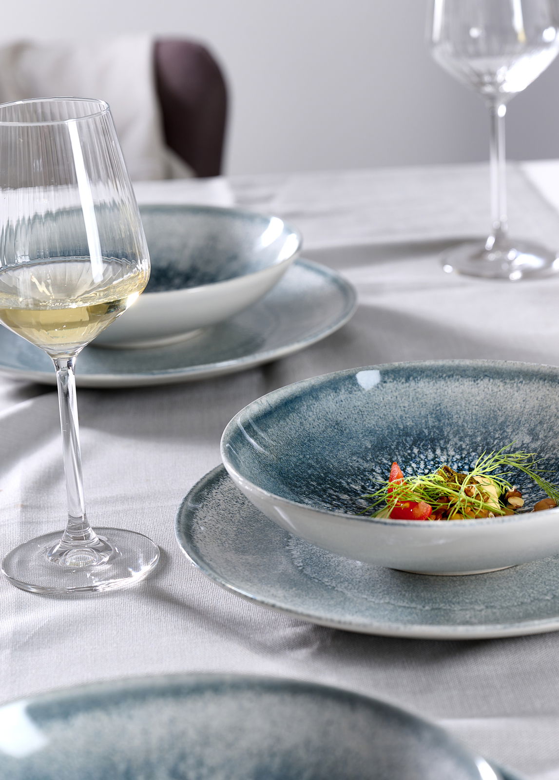 Nahaufnahme eines gedeckten Tisches mit einem eleganten Tellerset in Blau- und Grautönen, darauf ein kunstvoll angerichtetes Gericht, im Hintergrund ein Glas Weißwein.